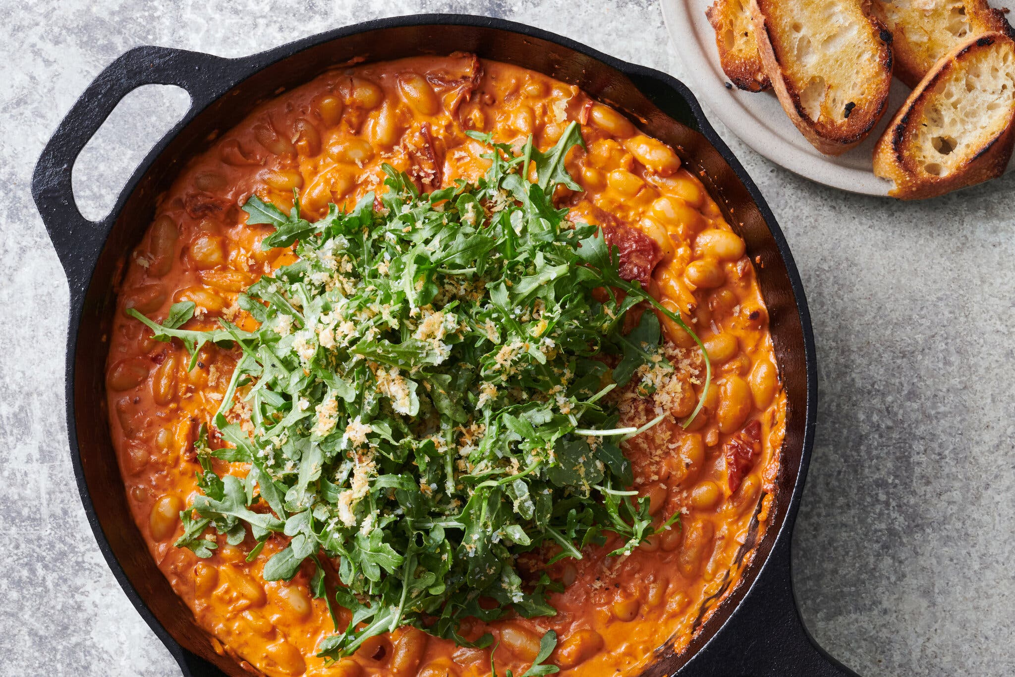 A cast-iron skillet is filled with beans in a creamy red sauce and topped with arugula that’s been tossed in bread crumbs.