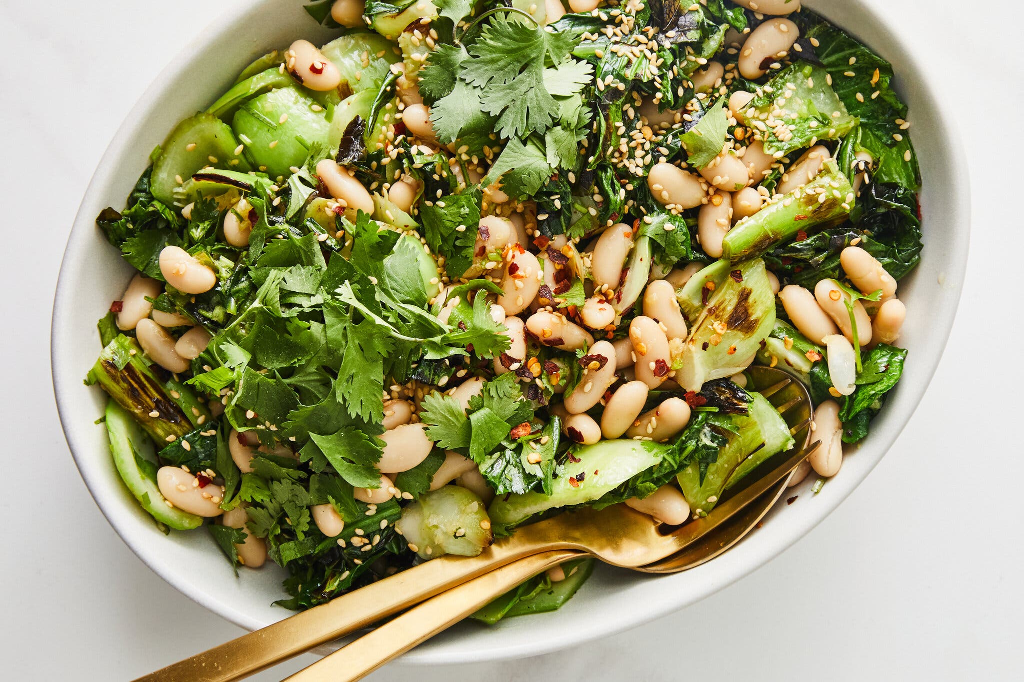 A white platter holds charred bok choy and cannellini bean salad with gold serving utensils.