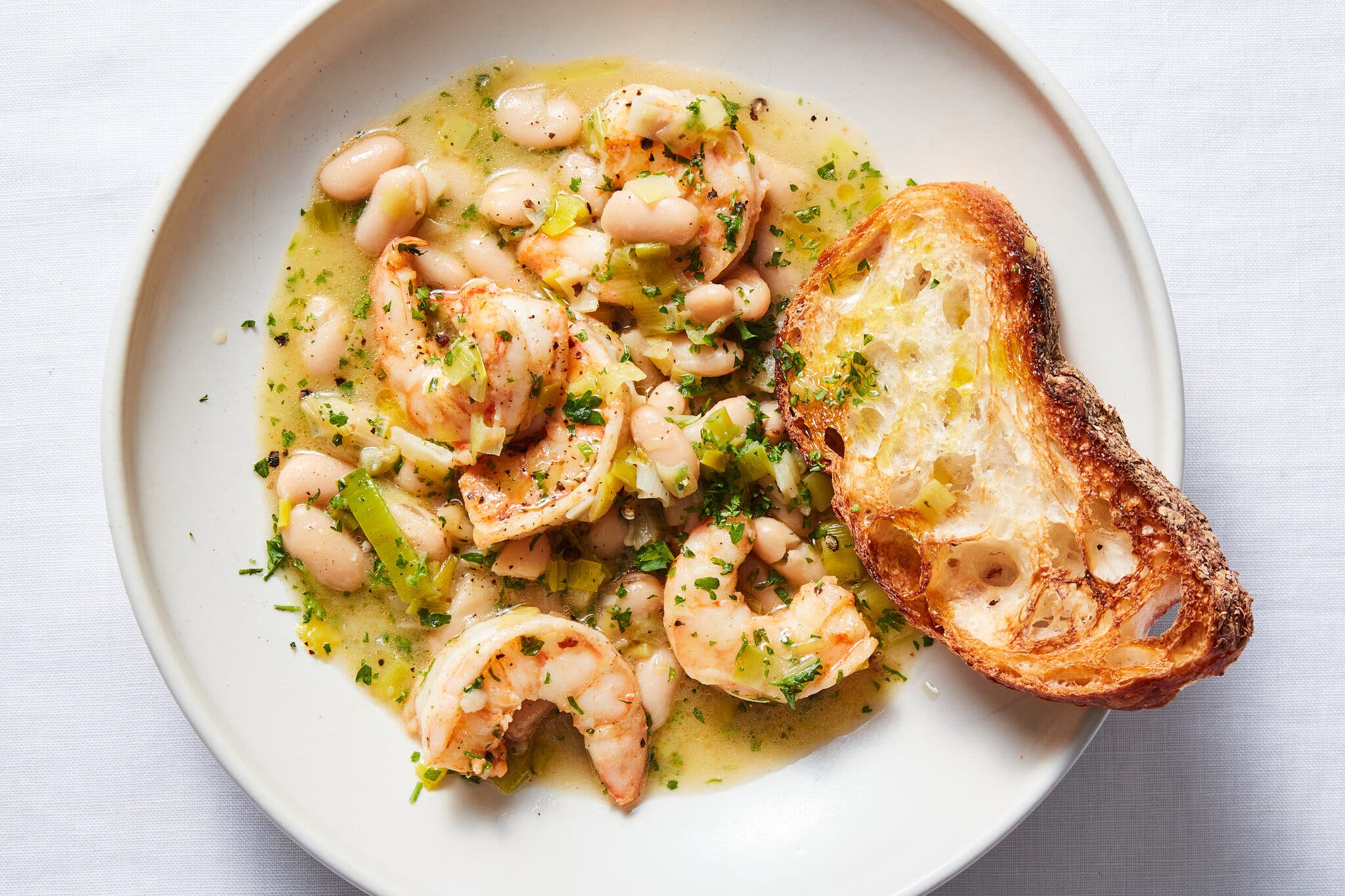 A rimmed white plate holds brothy beans and shrimp scattered with herbs and a piece of toasted bread.