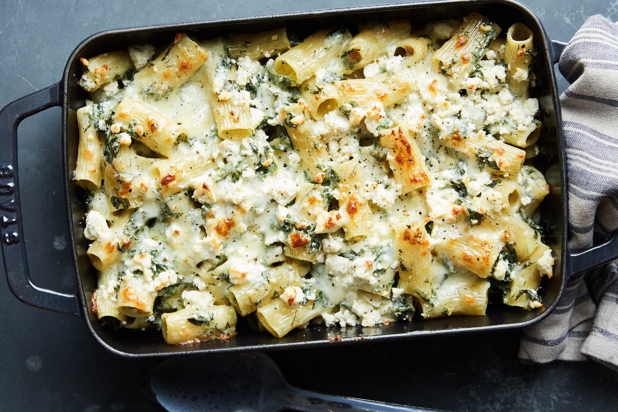 A baking dish filled with short pasta topped with cheese and mixed with greens.