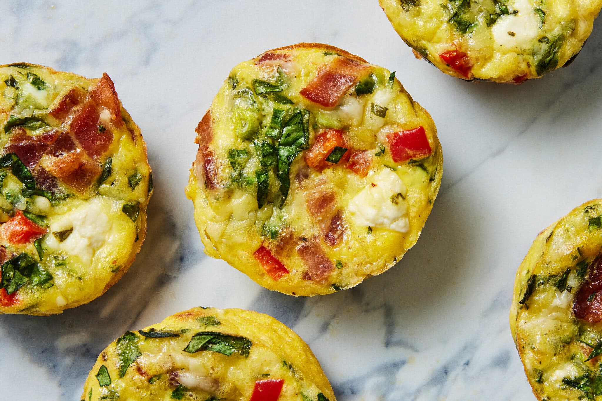 A close-up of egg muffins with bacon, spinach, and red pepper on a marble surface.