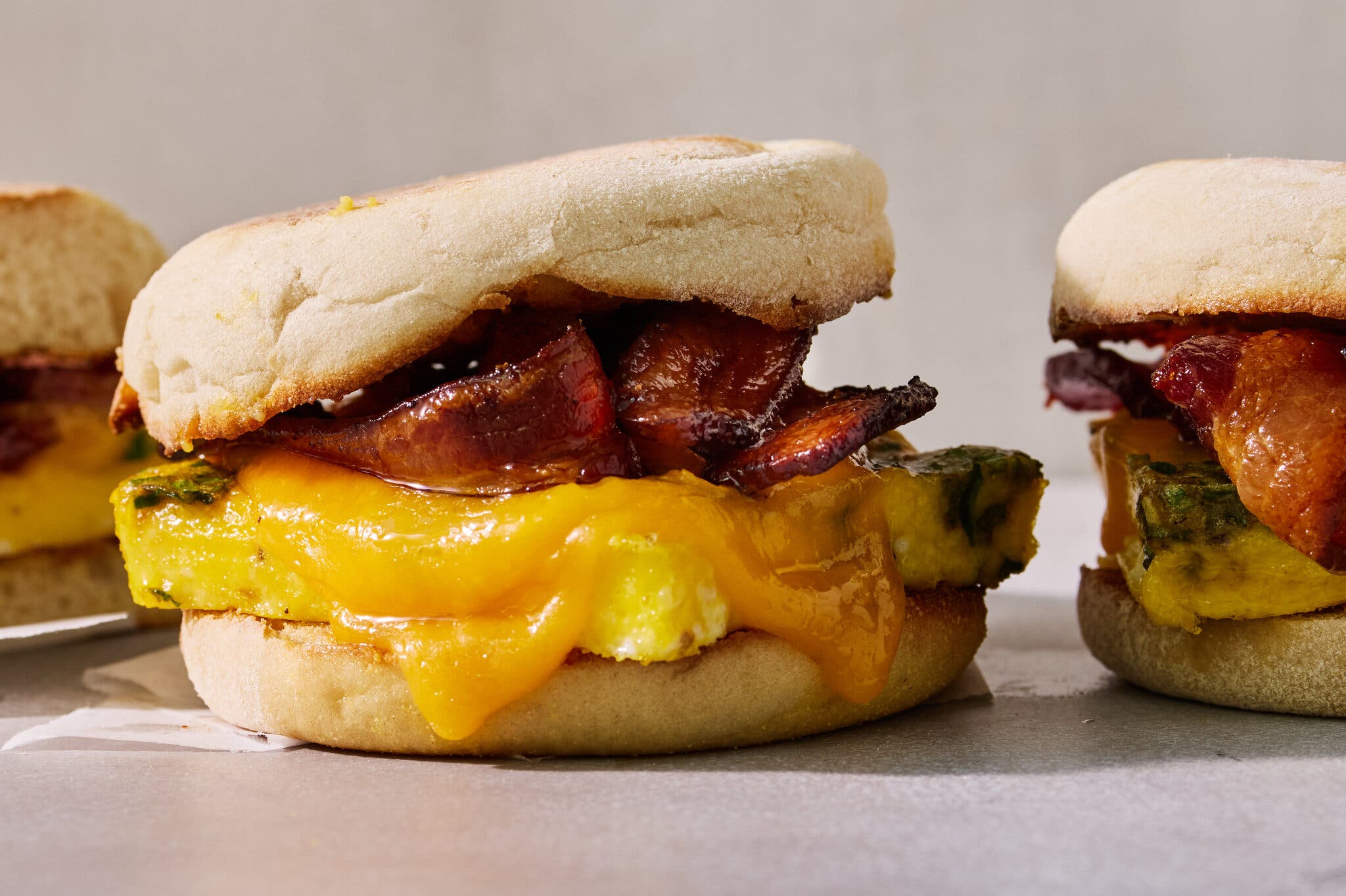 Three English muffin breakfast sandwiches with egg, cheese, and bacon on a white background.