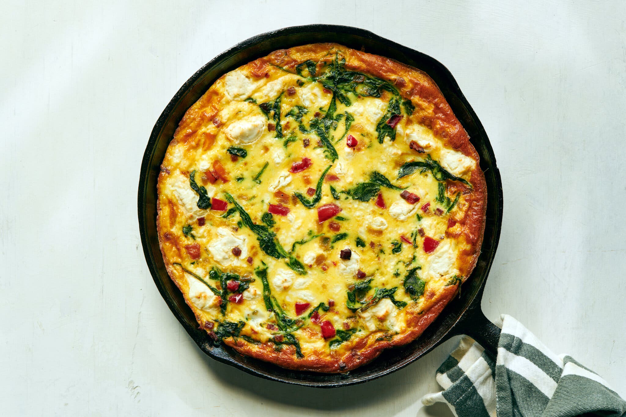 A cooked frittata in a black cast-iron skillet, with visible spinach, red peppers, and white cheese.