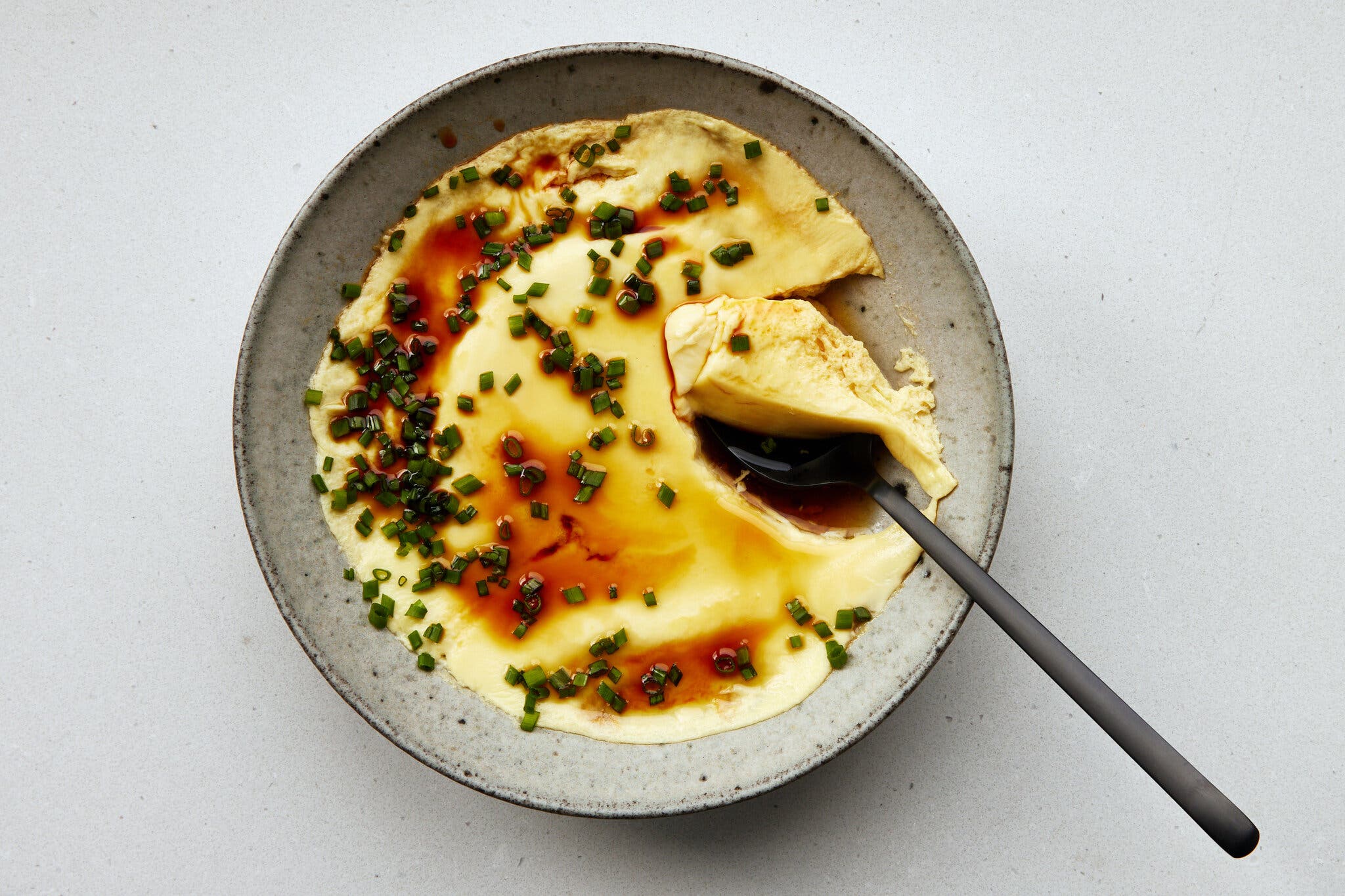 Steamed egg in a speckled bowl, topped with soy sauce and green onions, with a spoon scooping out a bite.