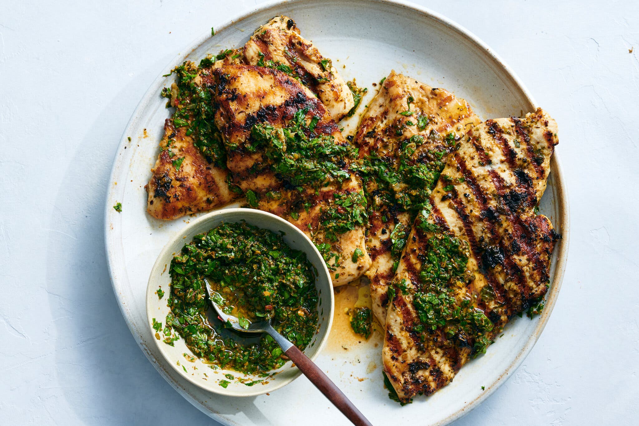 Pounded chicken breasts with sear marks sit on a plate with a bowl of verdant chimichurri.
