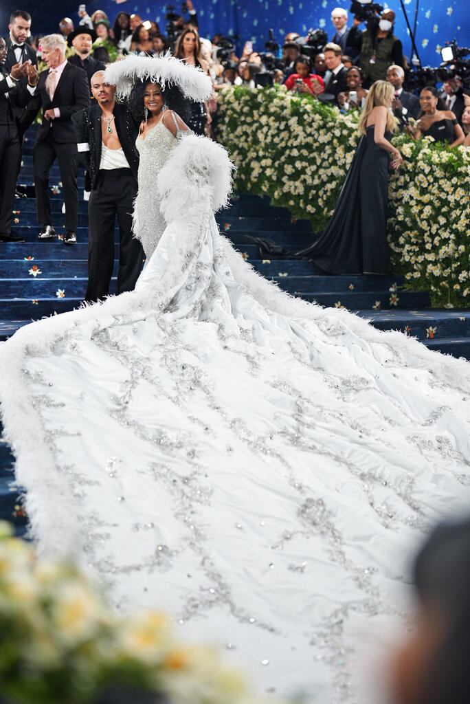 Diana Ross is wearing a silver dress, a wide-brimmed hat with feather details and a shawl with a long train.