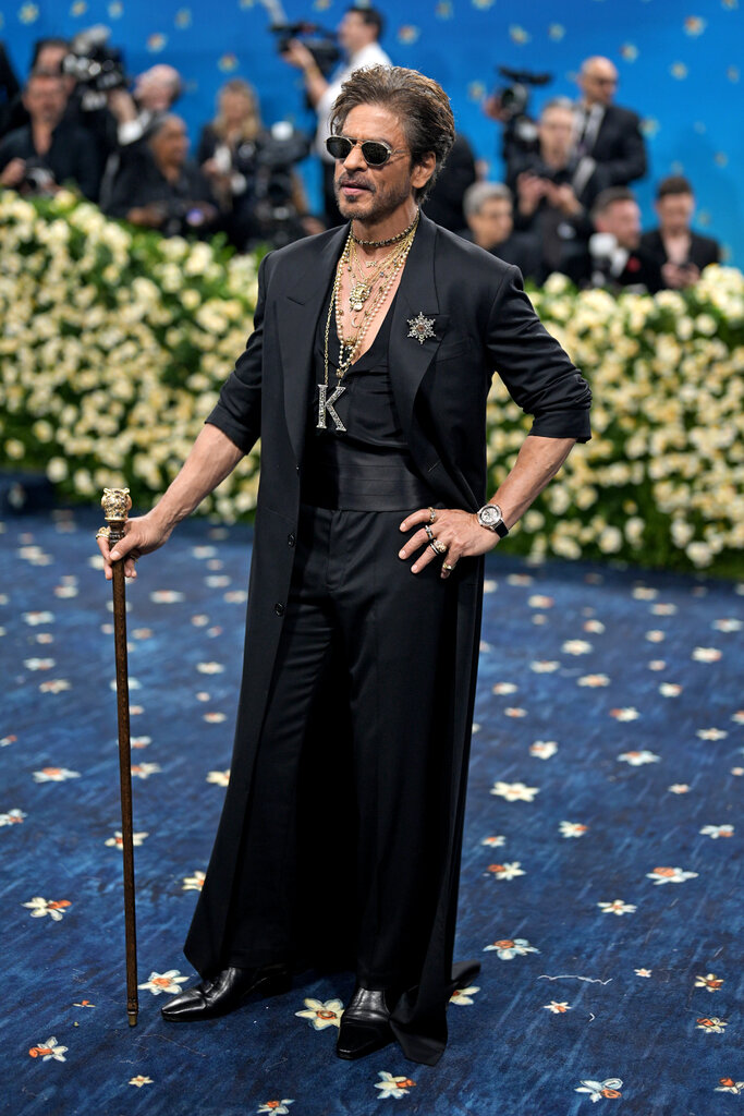 Shahrukh Khan is wearing a black suit and posing with a cane.
