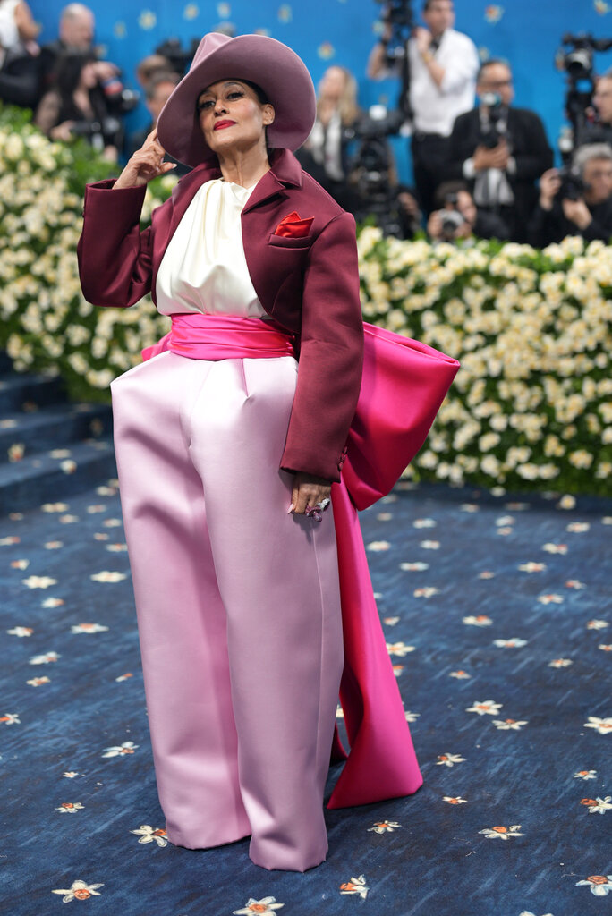 Tracee Ellis Ross wearing a cream blouse below a maroon suit jacket and pink wide-leg pants, with a hot pink oversize bow tied at the back and a wide-brim hat.
