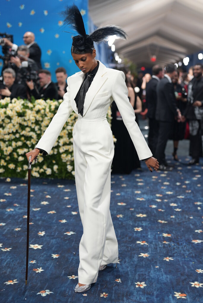 Imaan Hammam wearing a white suit and a hat adorned with feathers and posing with a cane.