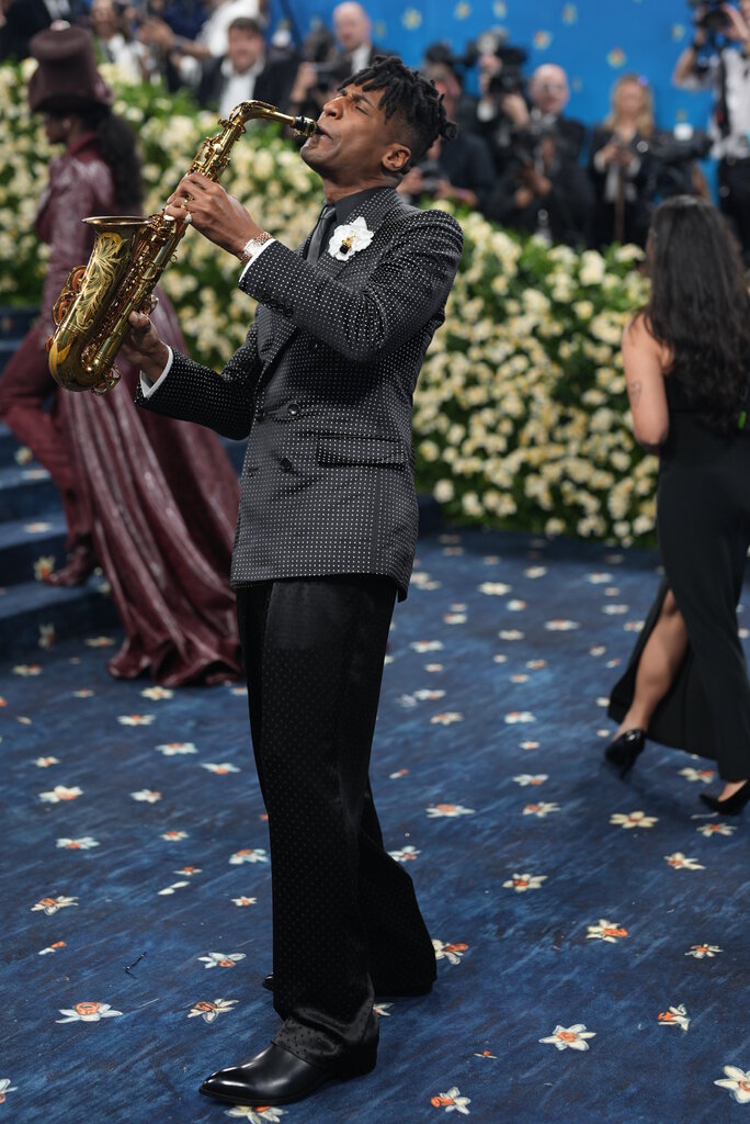 Jon Batiste is wearing a polka dot suit jacket and is playing the saxophone.