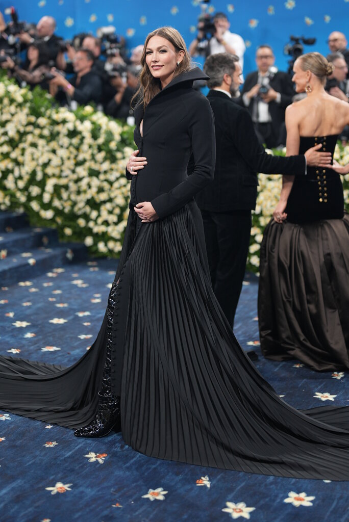 Karlie Kloss is wearing a black gown with a structured collar and pleated skirt.