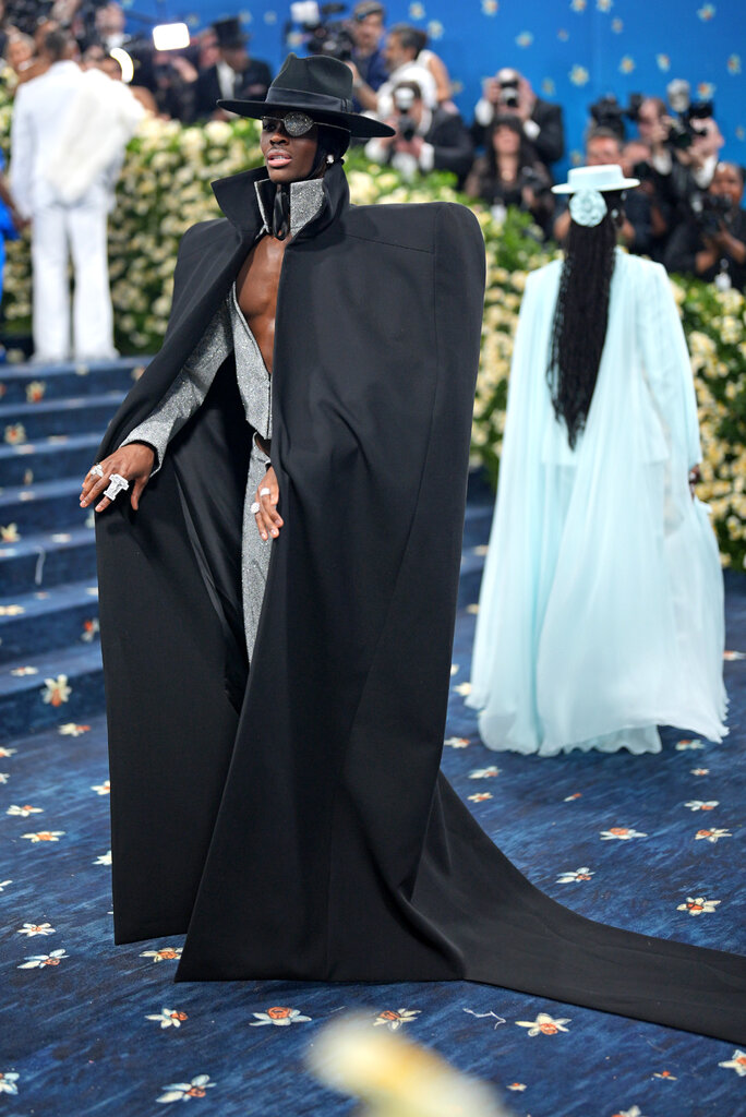 Alton Mason wearing a black floor-length cape with one exaggerated shoulder, a black wide-brim hat and silver eyepatch.