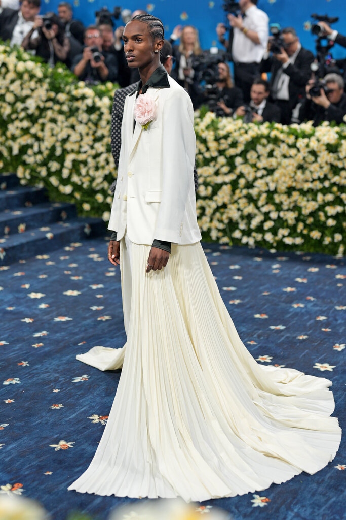 Christian Latchman wearing a cream colored blazer with a black satin collar, a pink flower brooch and floor-length train.