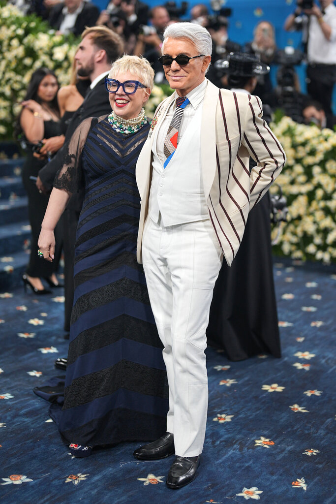 Catherine Martin, wearing a black and navy gown with lace detailing and blue-frame eyeglasses, poses with Baz Luhrmann, wearing a cream suit jacket with brown stripes overtop a white vest and trousers, a patchwork tie and black oval sunglasses.