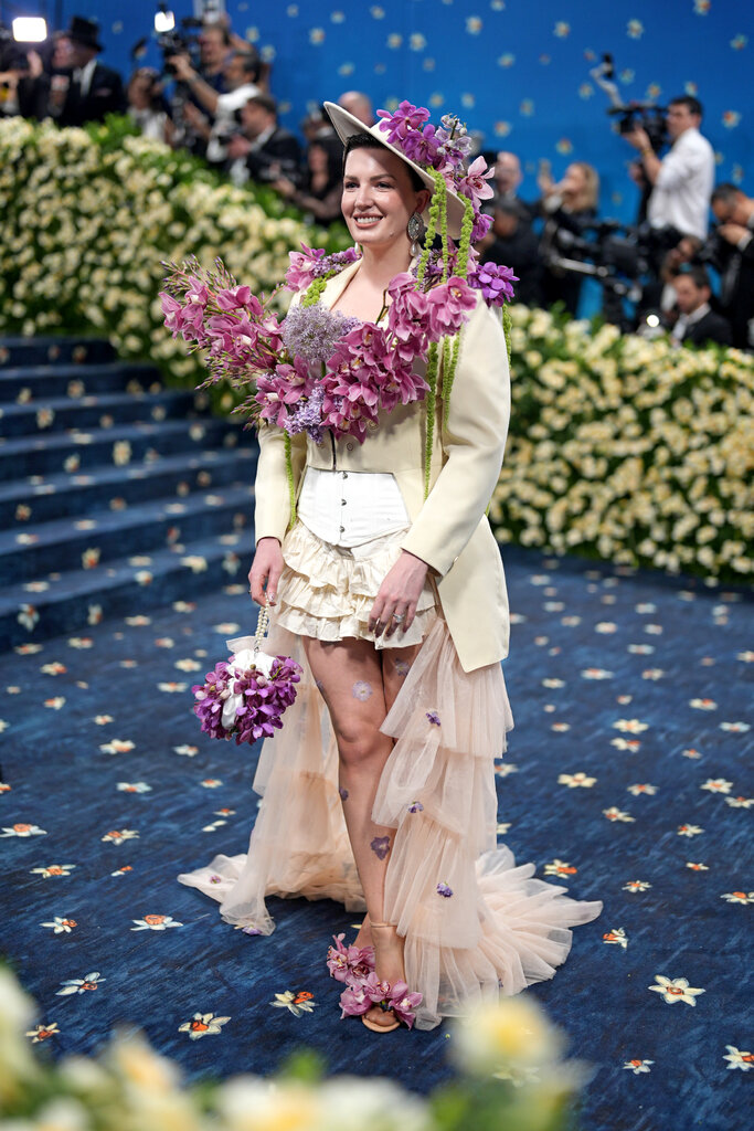 Jessica Kayll is wearing a creme suit jacket, corset, hat and ruffled skirt adorned with purple flowers.