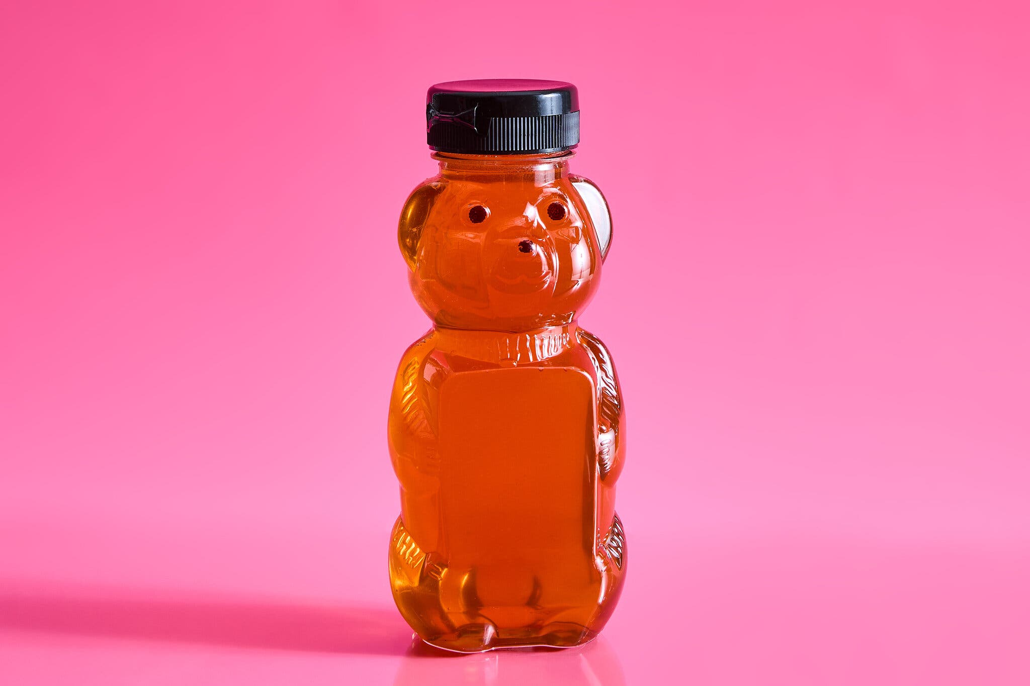 A bear-shaped plastic jar filled with honey sits against a hot pink background.