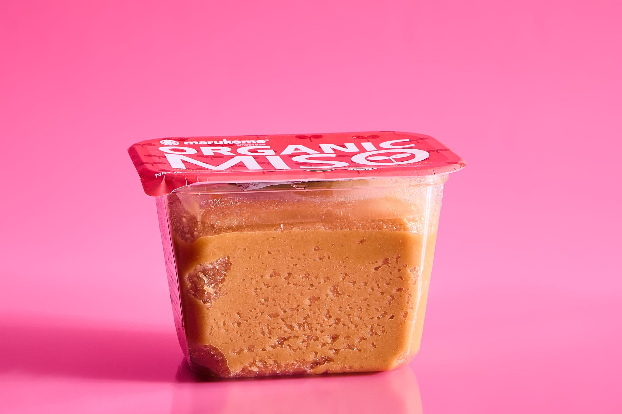 A tub of miso with a red lid against a hot pink background.