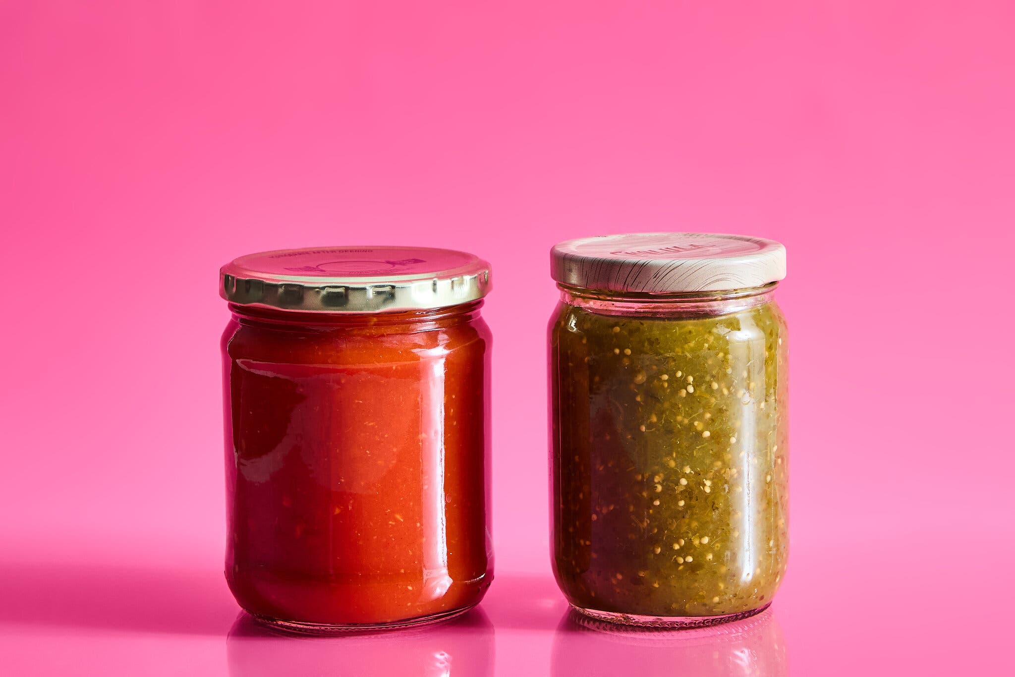 A jar of red salsa next to a jar of green salsa against a hot pink background.