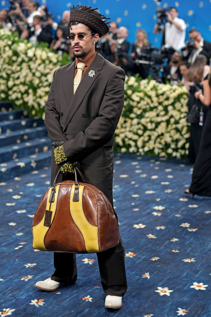 Bad Bunny wearing a brown suit, cream tie, brown straw hat. He is holding a large brown and yellow Prada bag.