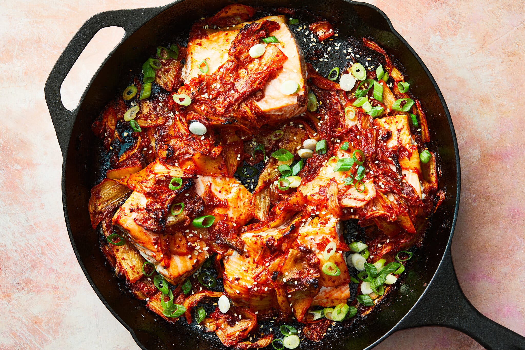 A cast-iron skillet holds fillets of salmon cooked with kimchi and garnished with green onion.
