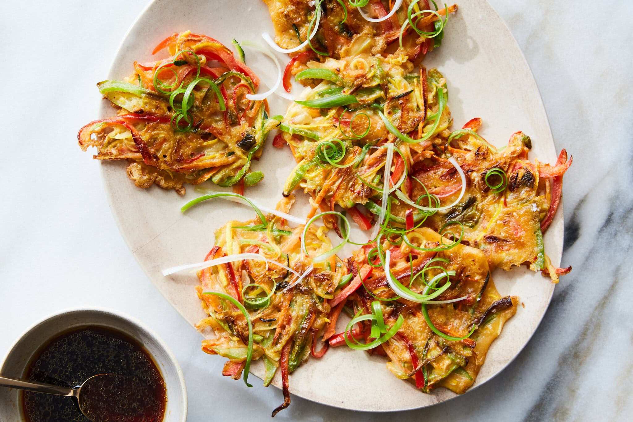 Several fried vegetable fritters sit on a plate. In the corner, there is a bowl of dipping sauce with a spoon in it.