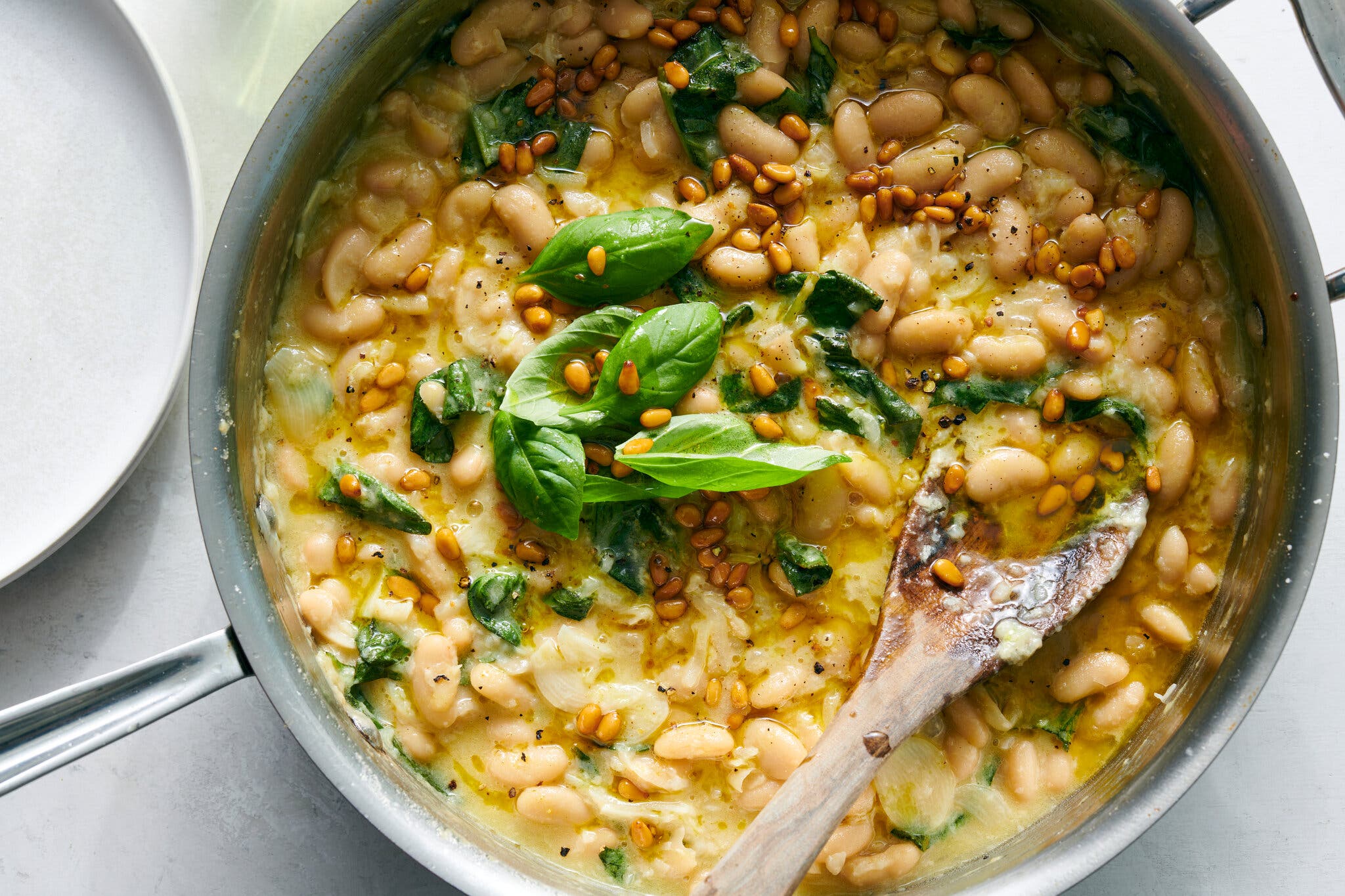 A skillet holds stewed cannellini beans garnished with fresh basil and pine nuts. A wooden spoon is scooping some of the beans.