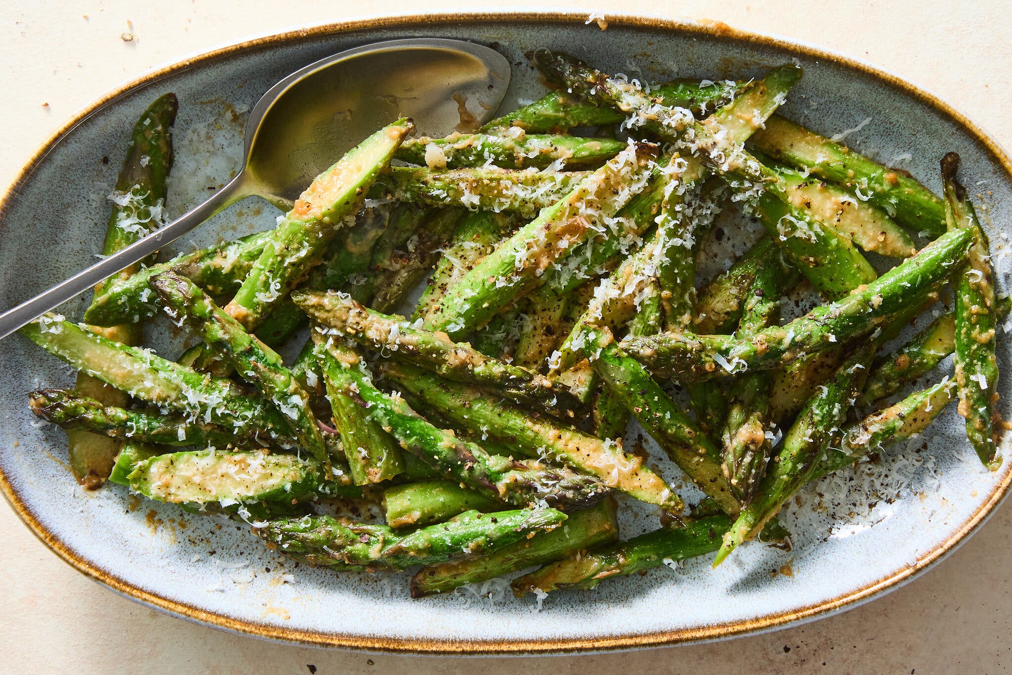 A long oval plate holds diagonally sliced asparagus, covered in miso sauce and garnished with grated Parmesan. There is a spoon coming into the frame.