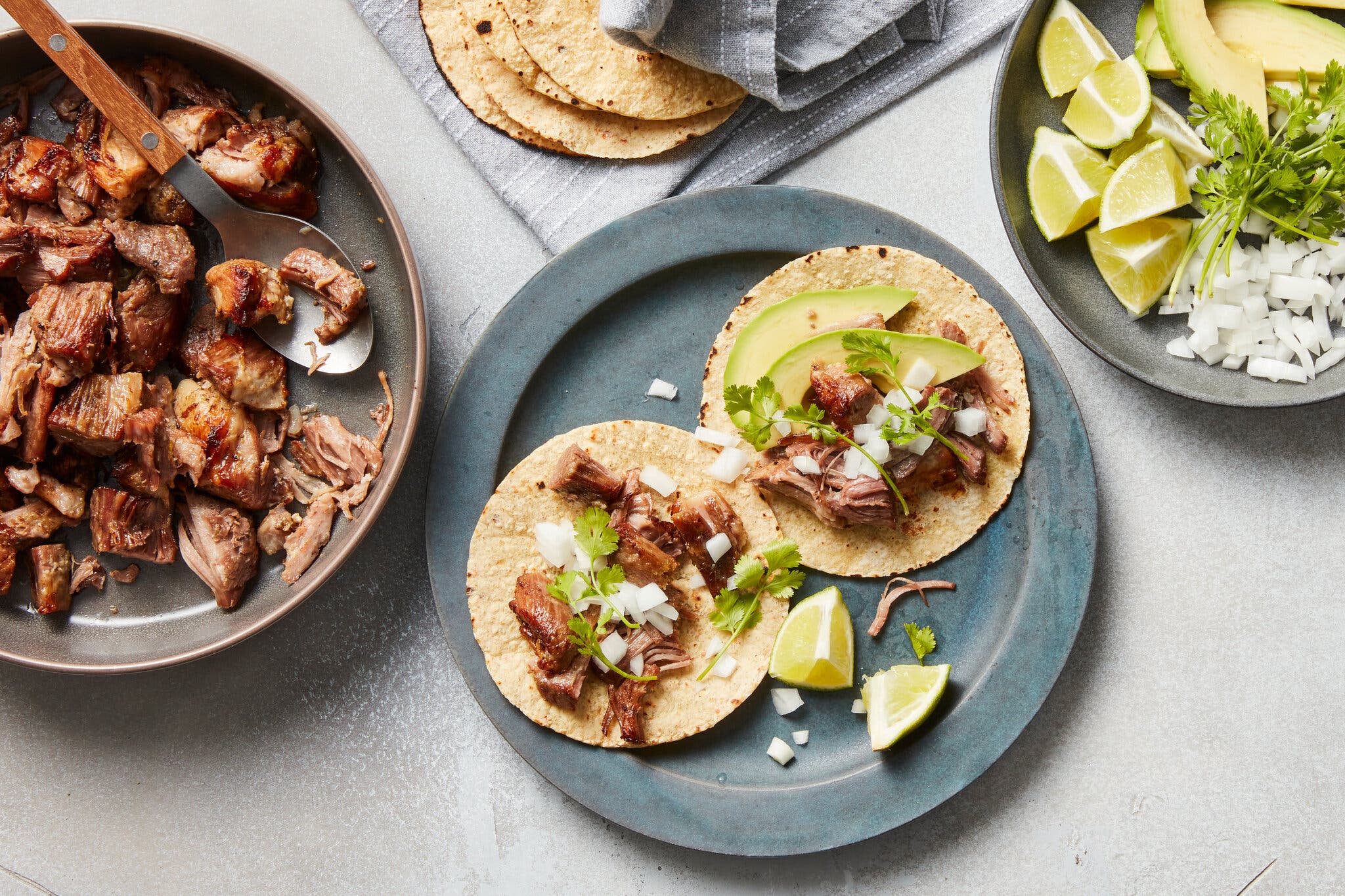 A blue plate holds two tortillas full of avocado, pork, diced onions and cilantro. There are lime slices on the plate. Surrounding the frame are a pan with pork, a plate of garnishes and some tortillas in a dish towel.