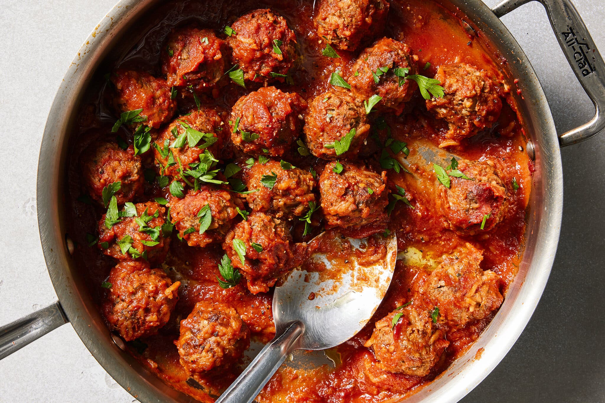 A large skillet holds meatballs covered in tomato sauce, garnished with herbs. A spoon is coming into the frame.
