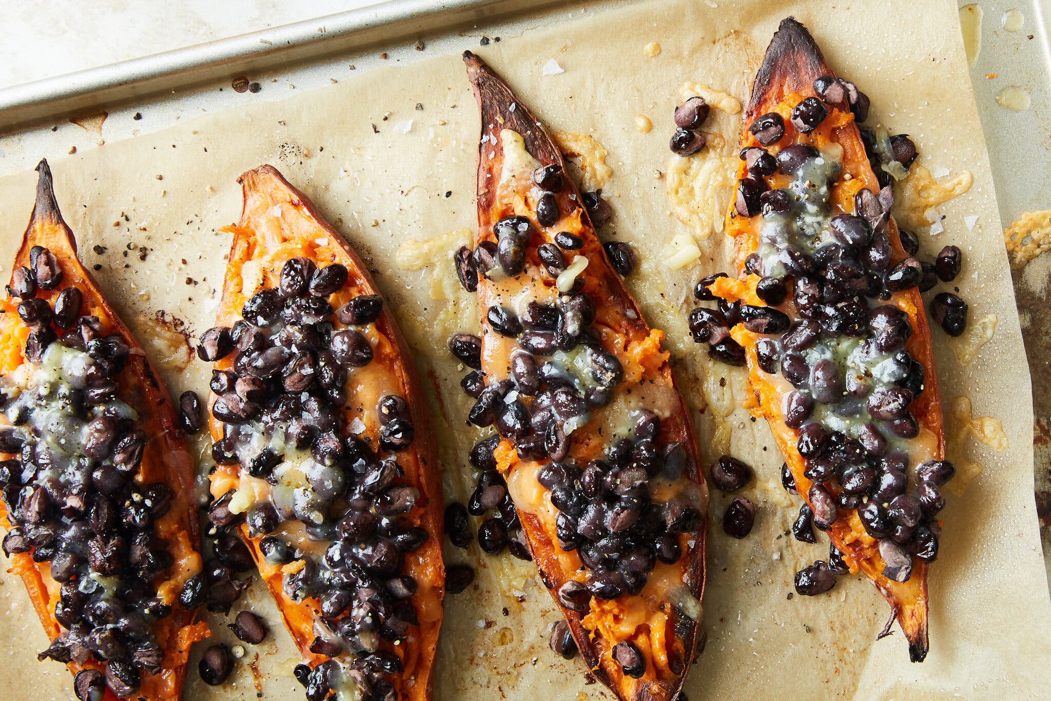 Four halved sweet potatoes lie on a baking sheet with parchment paper. They are topped with melted cheese and black beans.