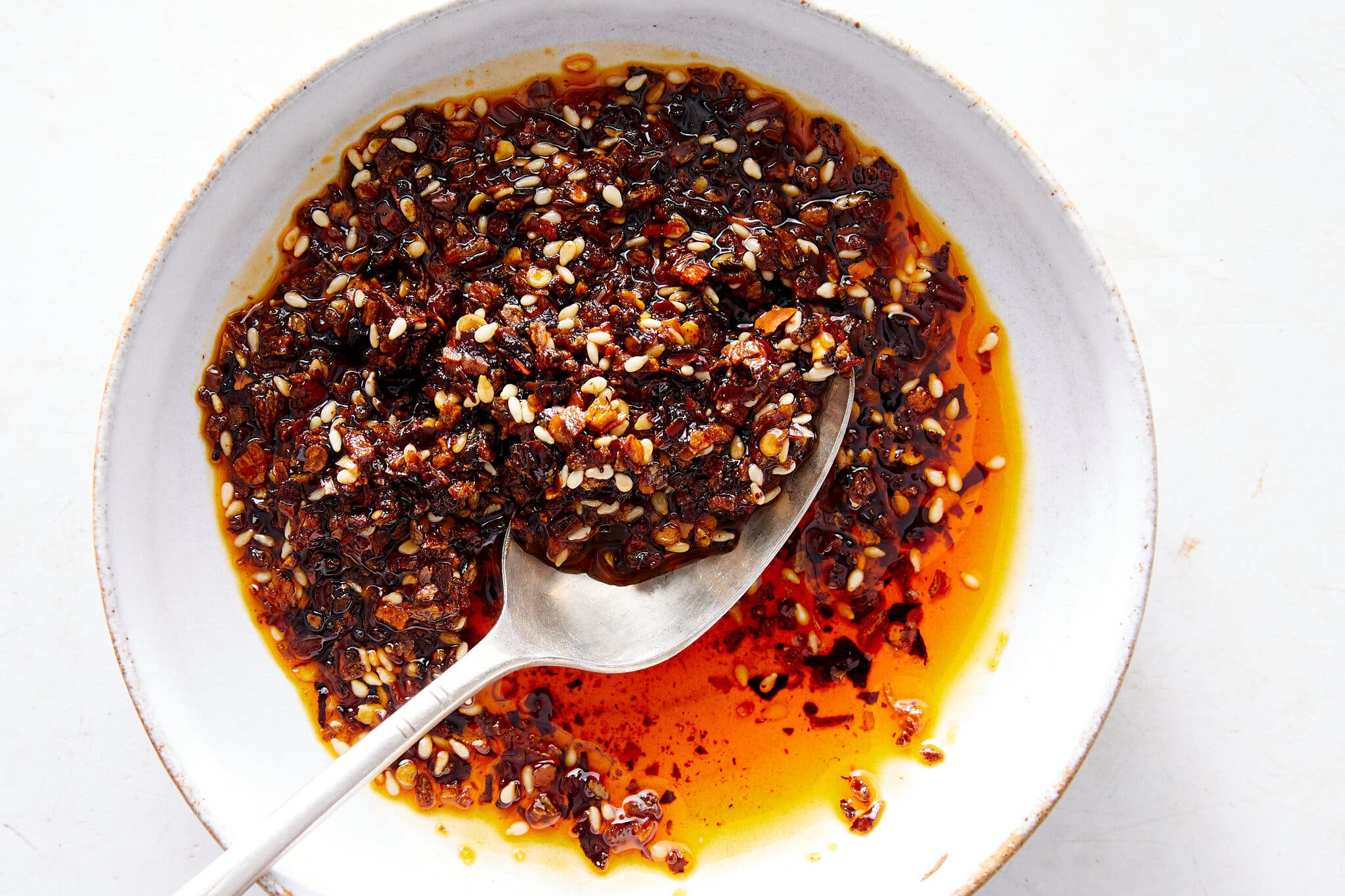 A bowl holds chile flakes and sesame seeds in bright red oil. There is a spoon scooping some of the chile crisp.