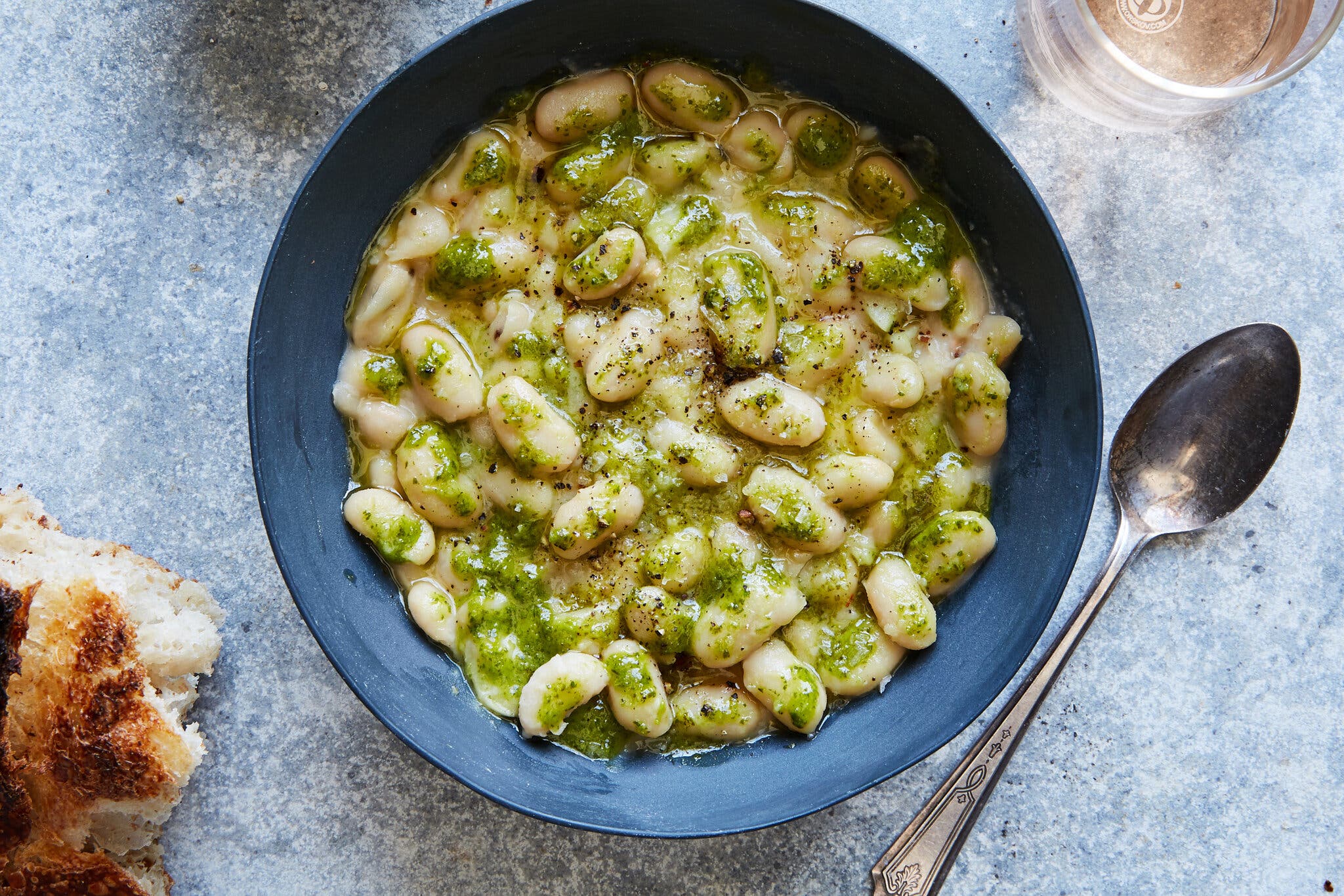 A blue bowl holds cannellini beans garnished with green oil and black pepper. A piece of bread and a spoon surround it.
