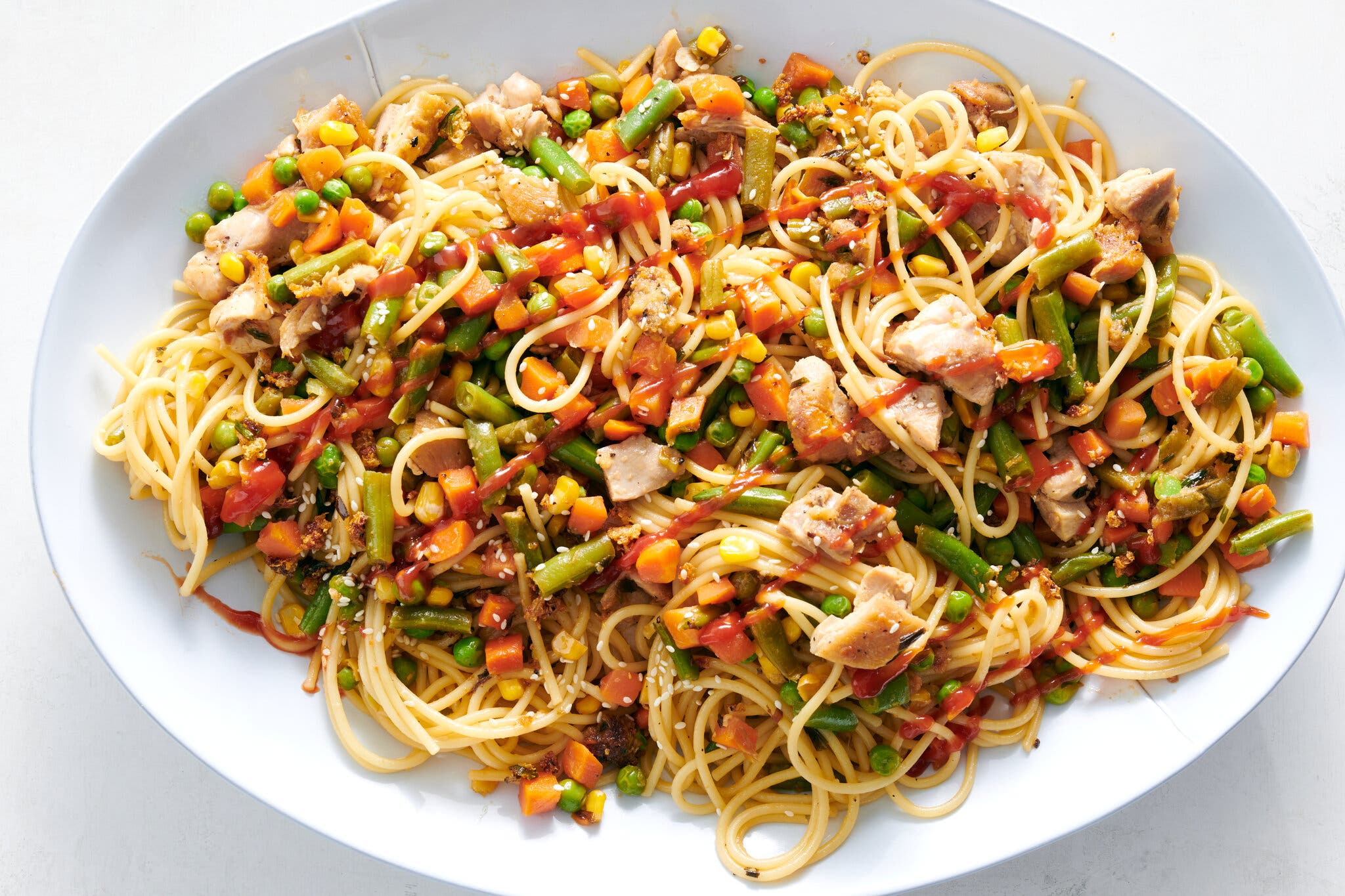 Photographed from above: A large white oval dish with chicken, spaghetti, carrots, peas, string beans, drizzled with red sauce.