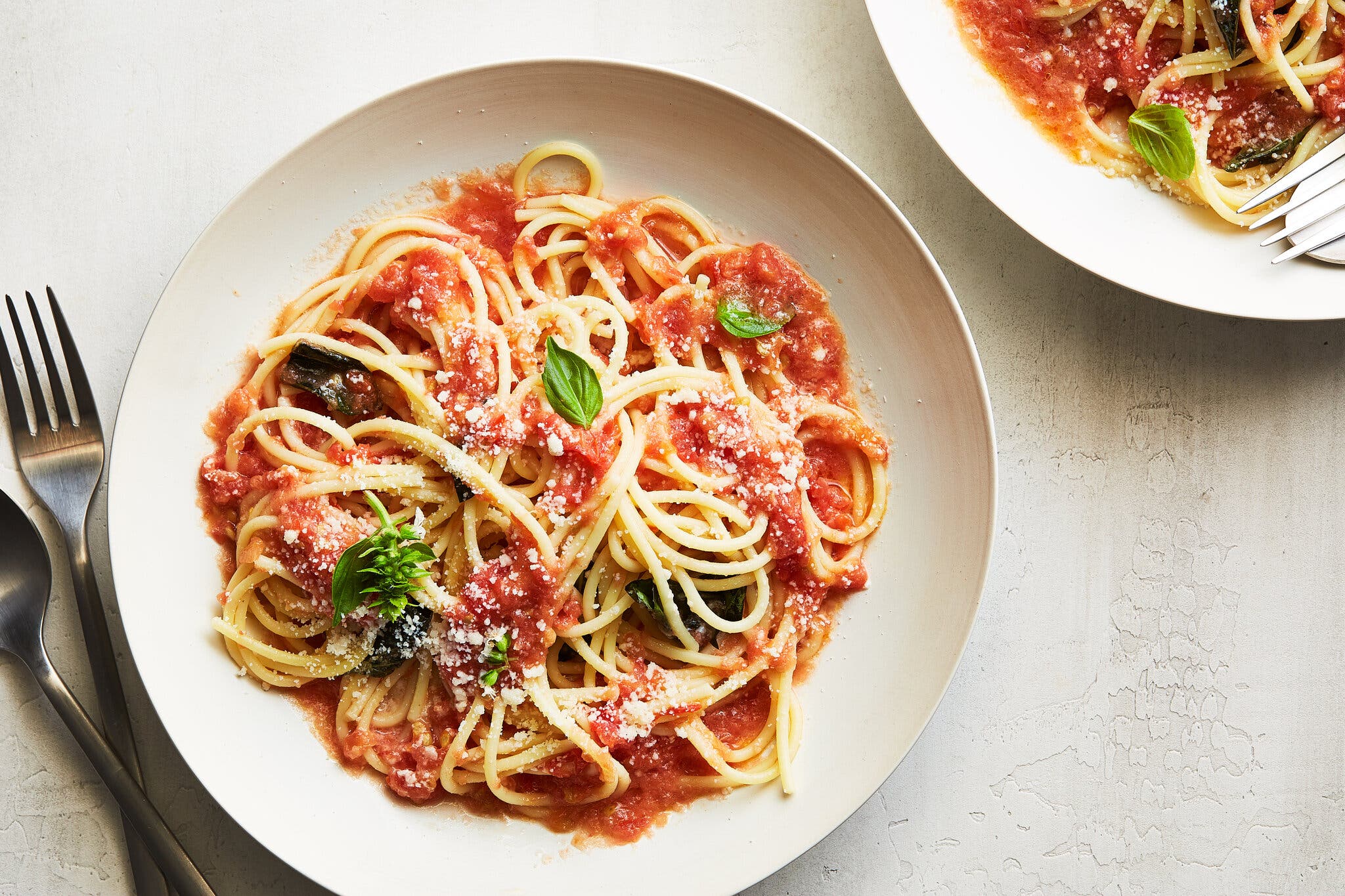 Photographed from above: a bowl of spaghetti with tomato sauce, garnished with basil. To the left sits a spoon and fork.