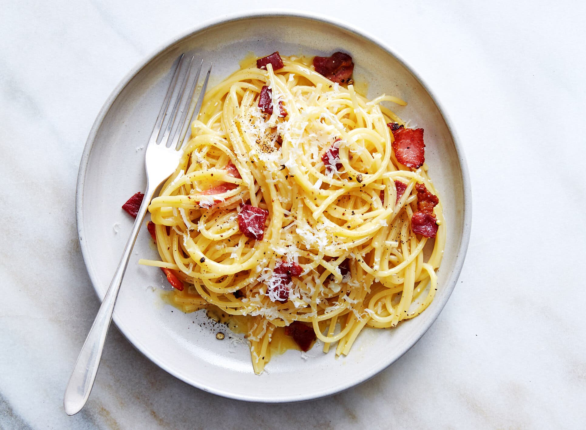 A bowl of spaghetti carbonara on a white plate sits on a gray-and-white marble countertop.