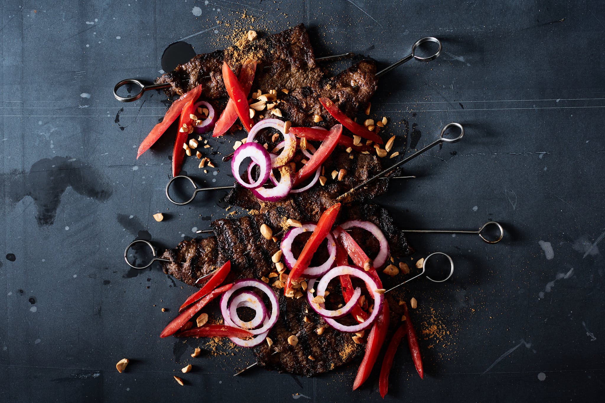 Skewered beef topped with red onions and red peppers on a dark background.