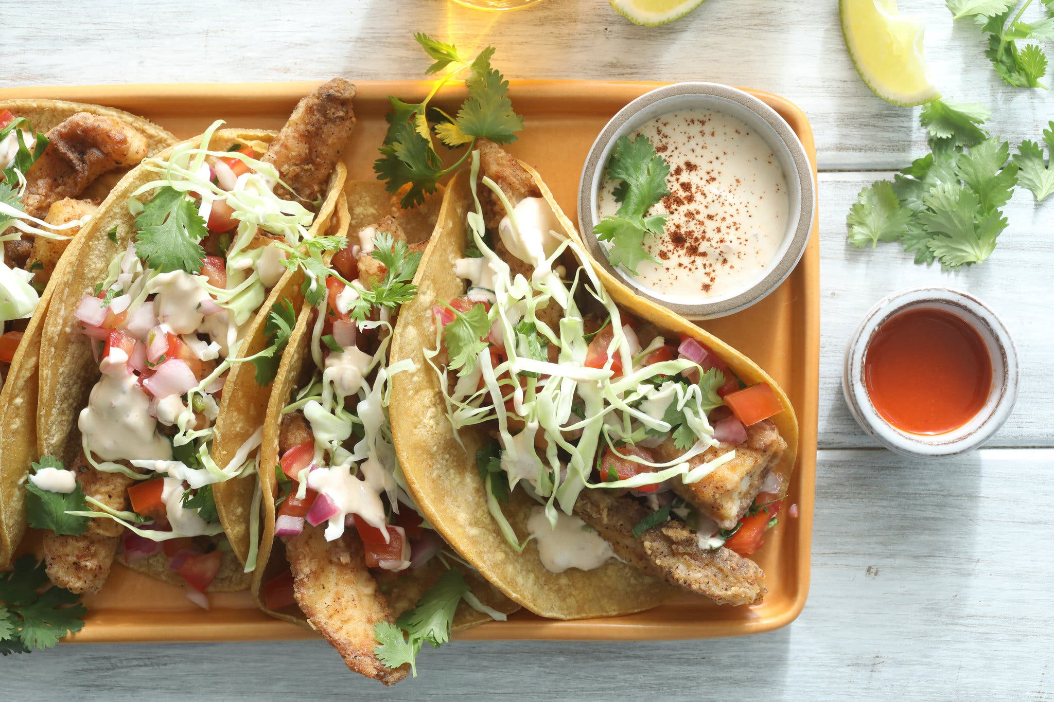 An overhead shot of a wooden tray holding three fish tacos, garnished with crema and cilantro. In the top left corner of the tray is a dish of white sauce. To the right of the photo is a dish of hot sauce.