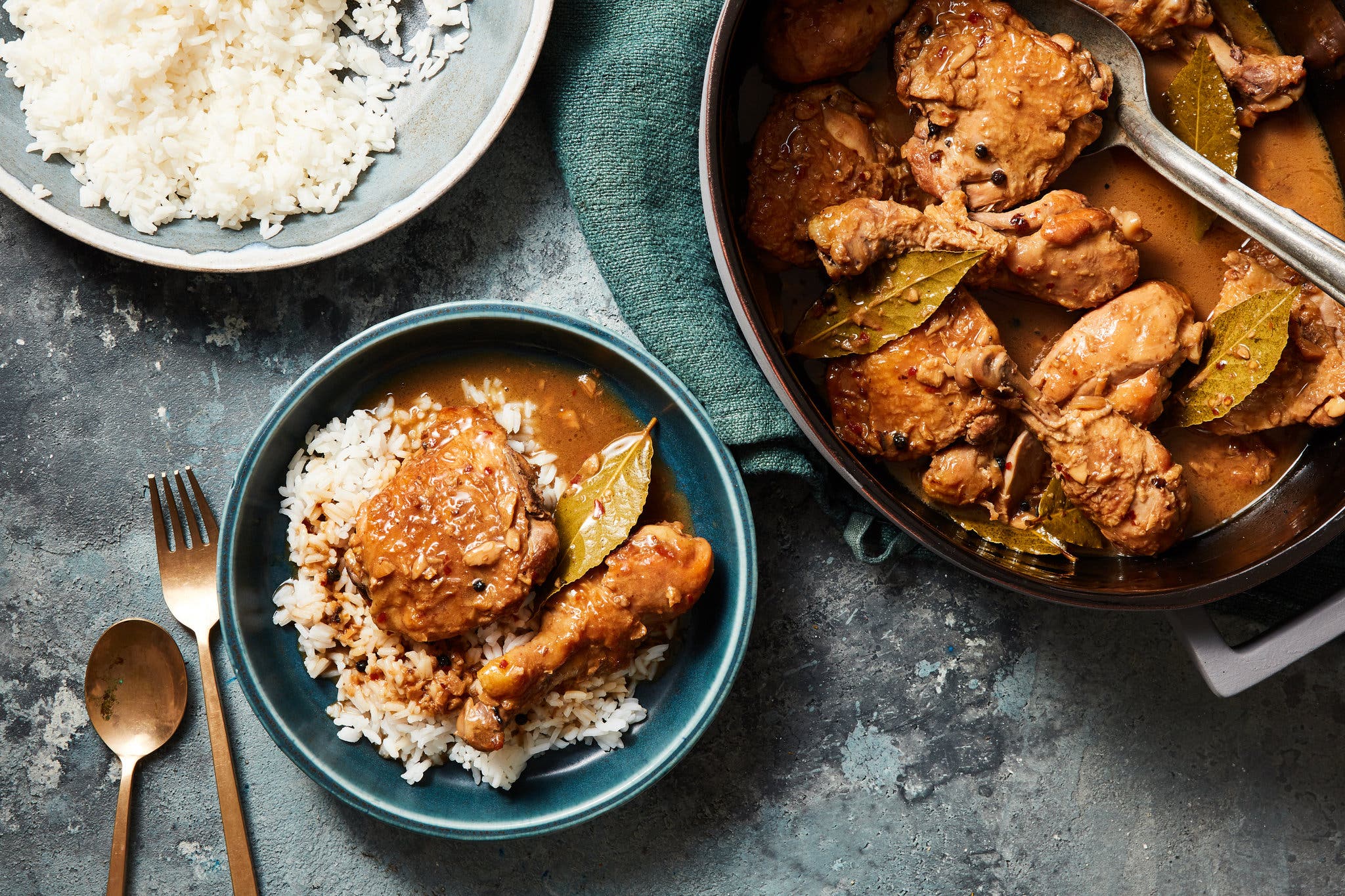overhead shot of a blue bowl of chicken adobo, garnished with a bay leaf. A large pot with serving spoon sits to its right.
