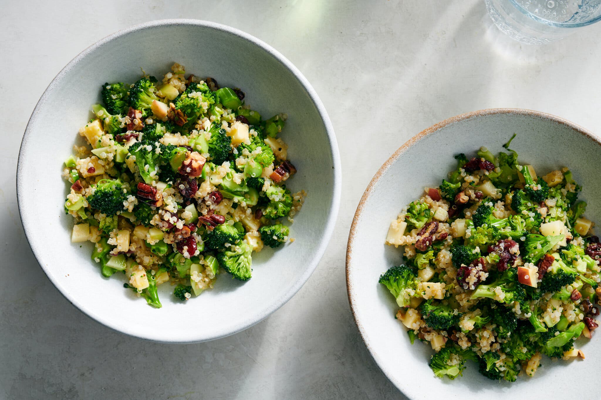 Two light bowls filled with broccoli, quinoa, apples and nuts are photographed from overhead.