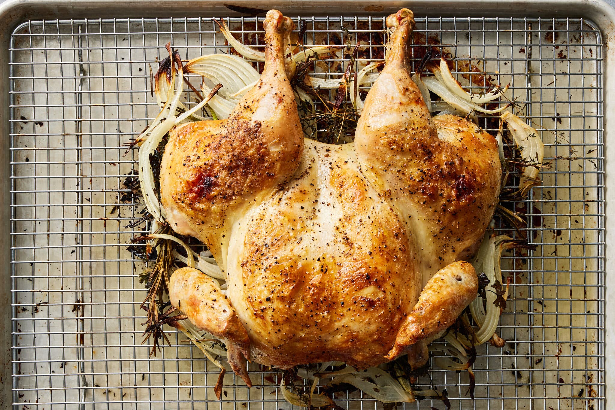 A spatchcocked roast chicken is shown on top of burnished onion slices on a cooling rack set inside a baking sheet.