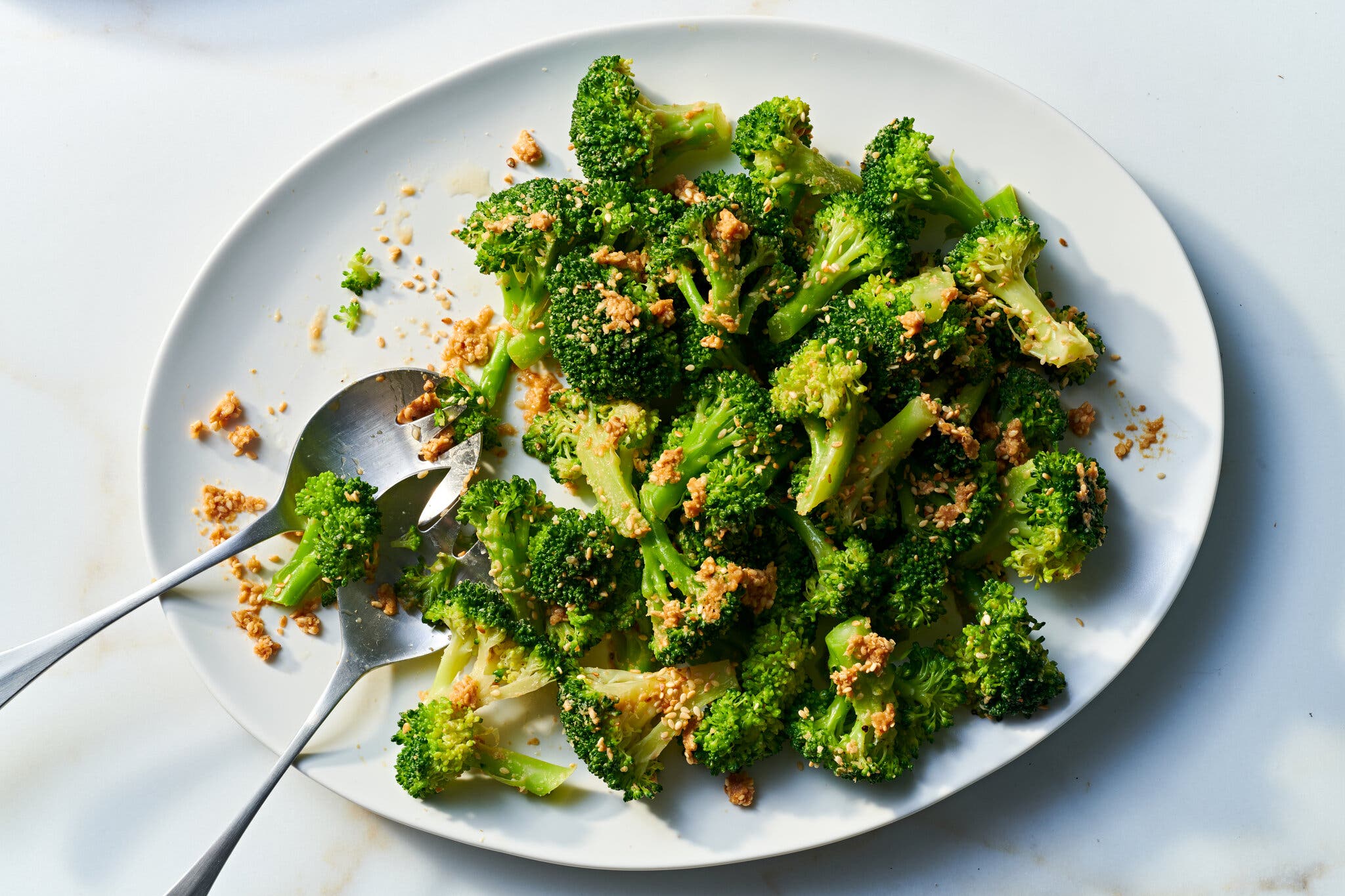 photographed from above: a large white oval serving dish with broccoli, garnished with sesame seeds. a serving spoon and fork are on the dish.
