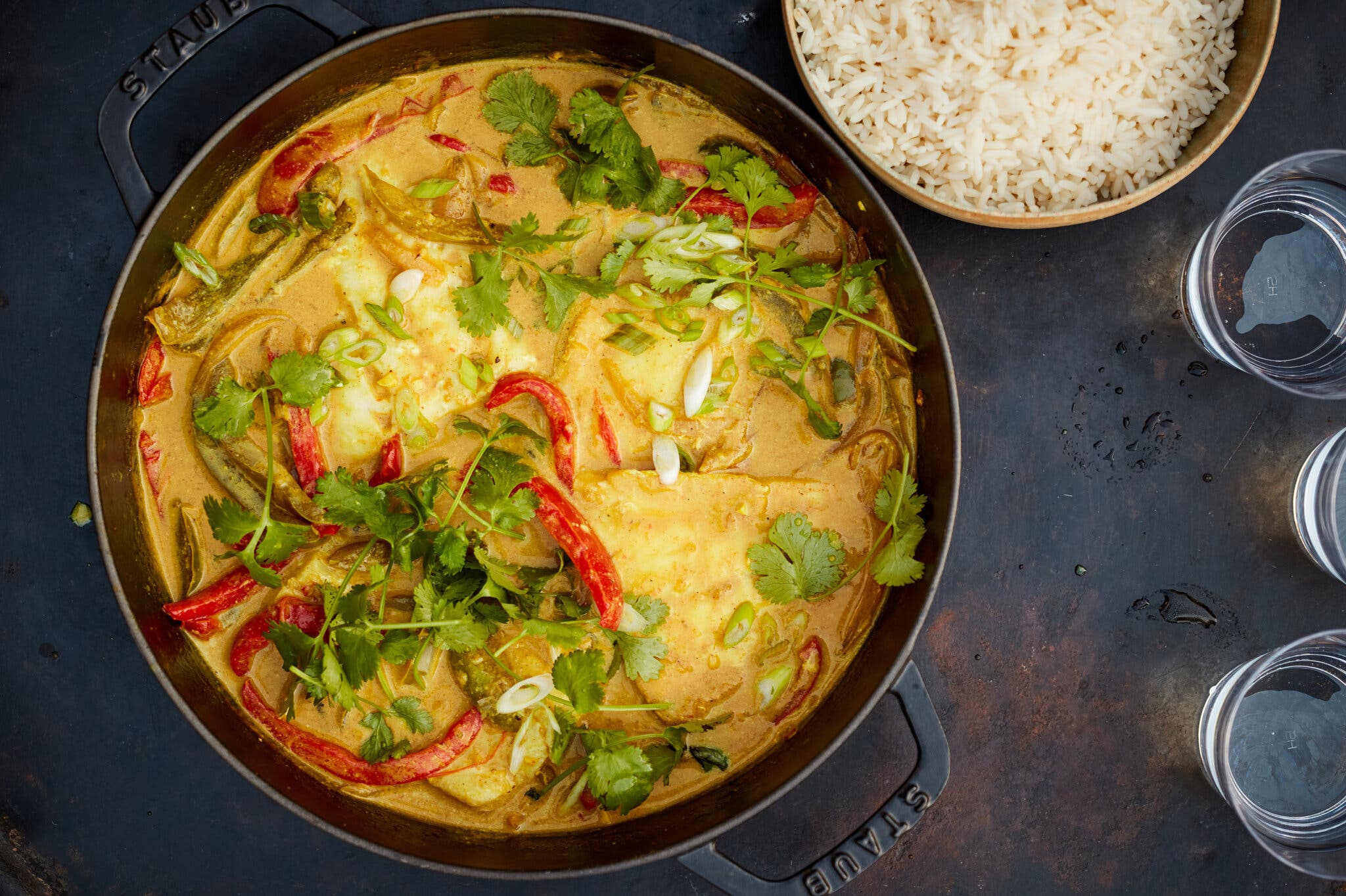 A Dutch oven holds a colorful coconut fish curry with red peppers and green cilantro sprigs. A bowl of rice is nearby.