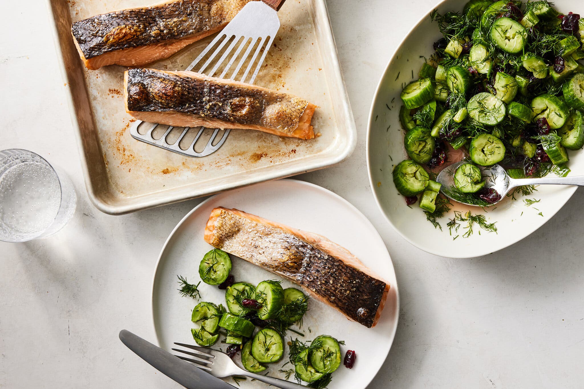 An overhead shot shows roasted salmon with crispy skin on a baking sheet and a dill-cucumber salad in a white bowl.