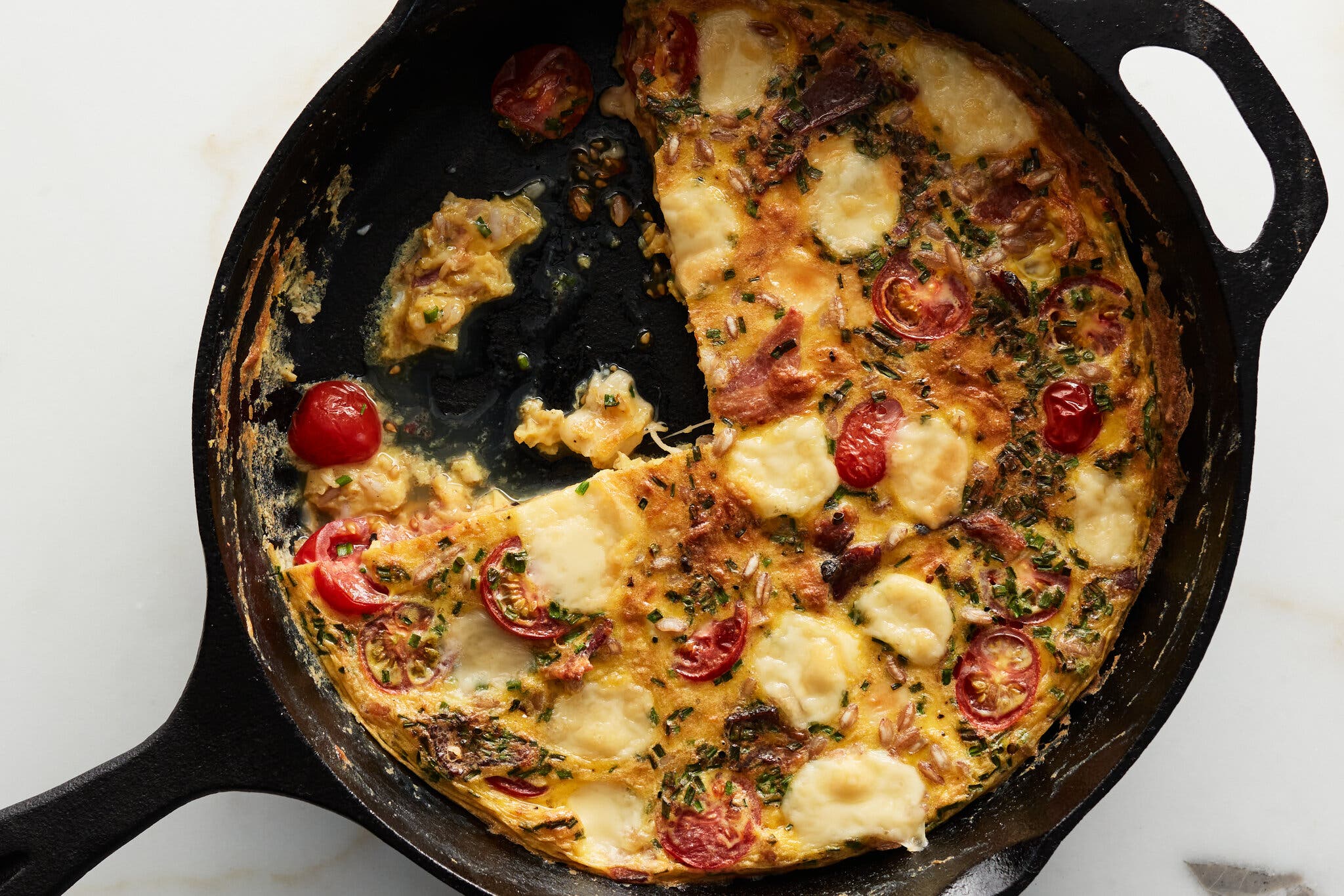 A cast-iron skillet holds a cheesy frittata with cherry tomatoes and dollops of cheese.