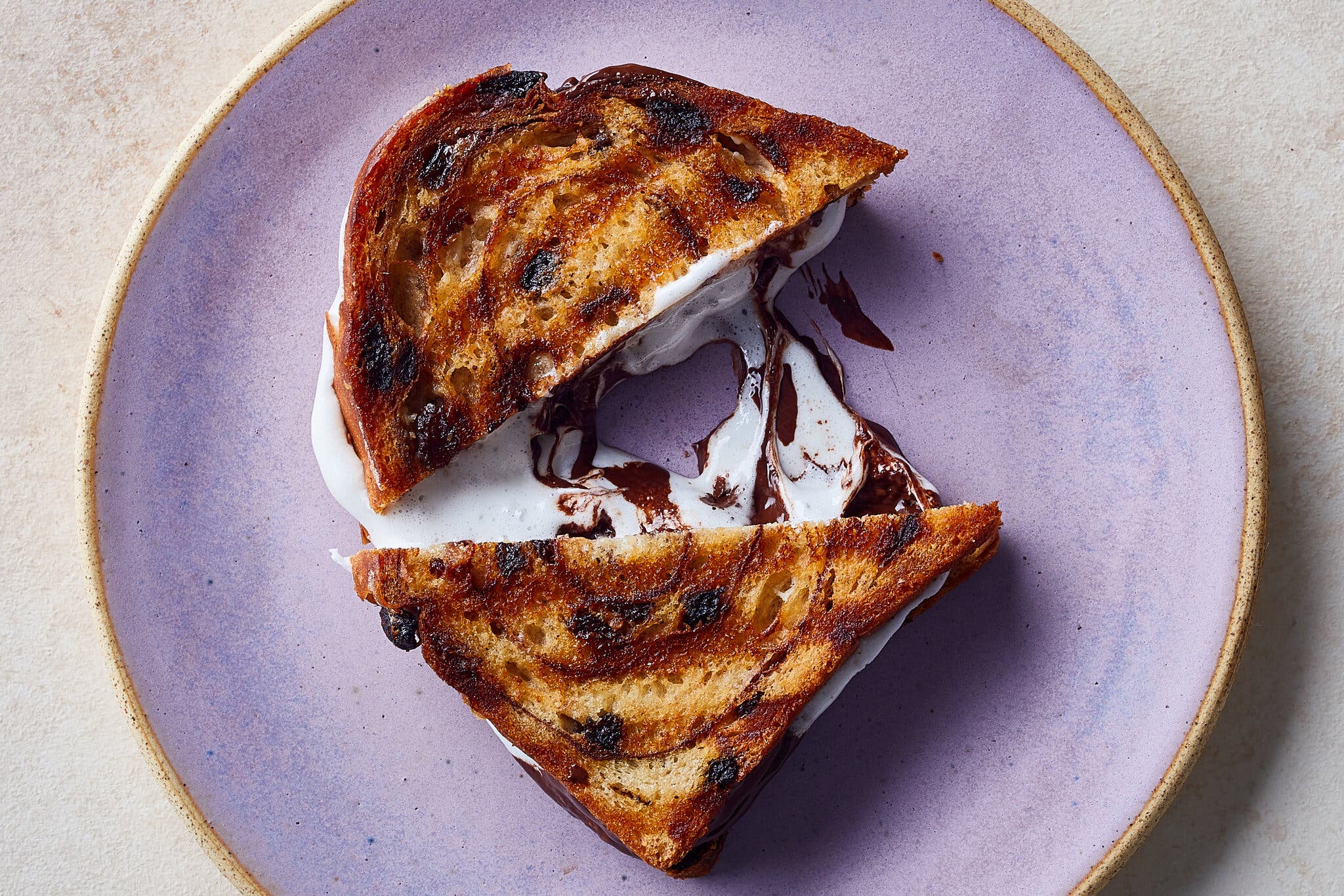 A grilled s’mores sandwich on cinnamon swirl bread with raisins sits on a purple plate. The sandwich is cut diagonally in half, with the chocolate and marshmallow filling oozing out.