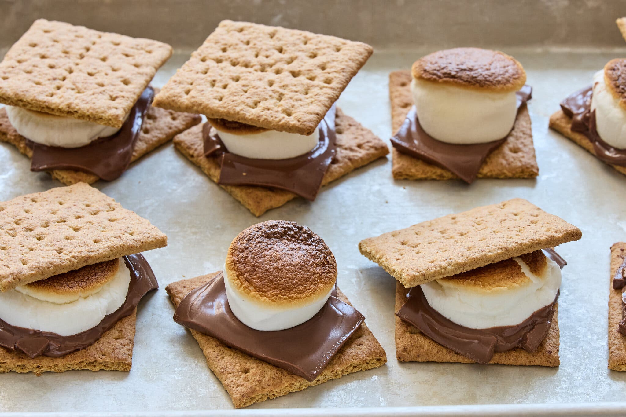 Six s’mores on a light gray surface. Two are not topped with graham crackers, showcasing the golden brown exterior of the toasted marshmallows.
