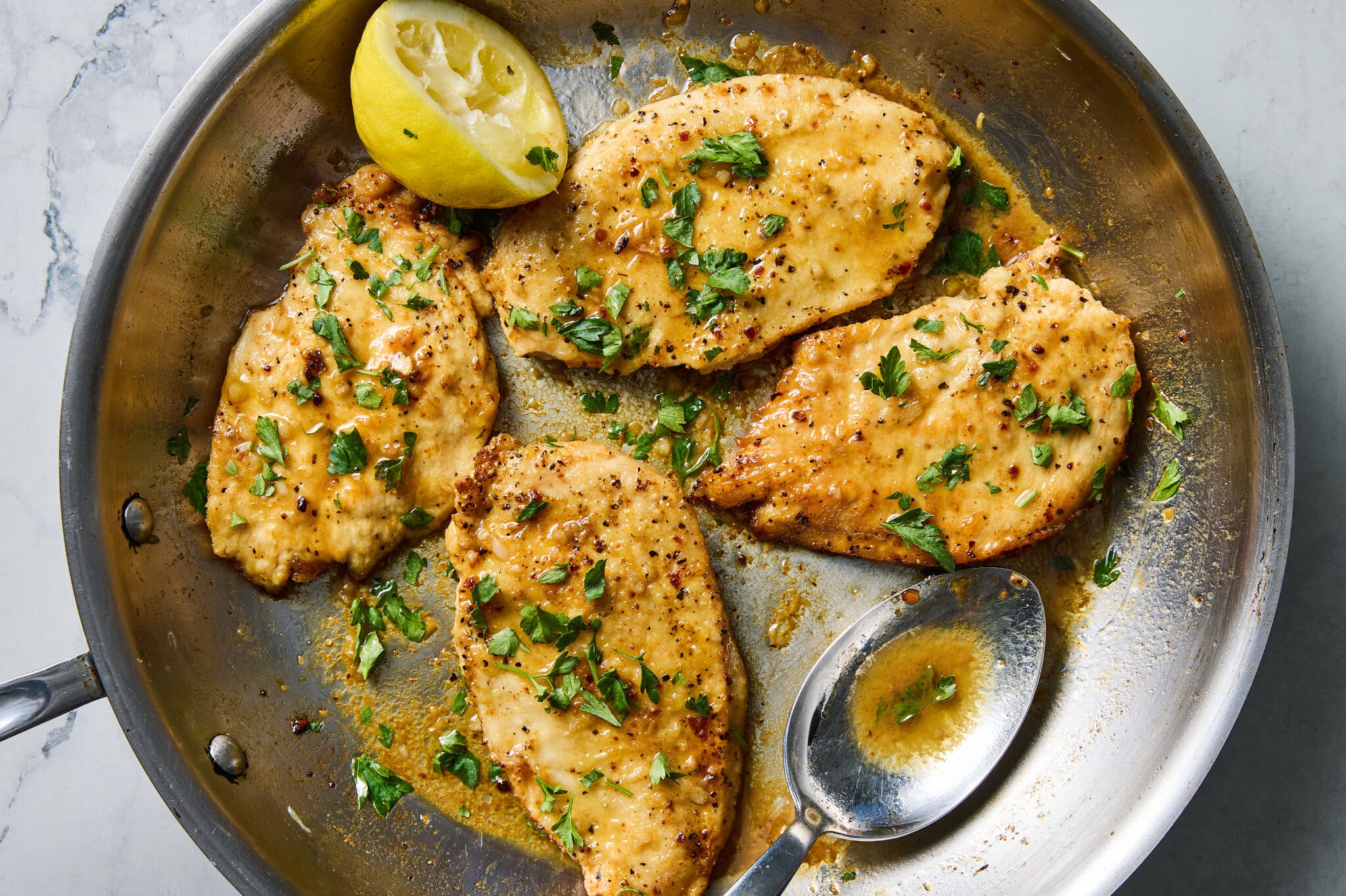 An overhead image of four cooked boneless skinless chicken breasts in a skillet. They’re glossed with a lemony sauce and sit next to a spoon and a spent lemon half.