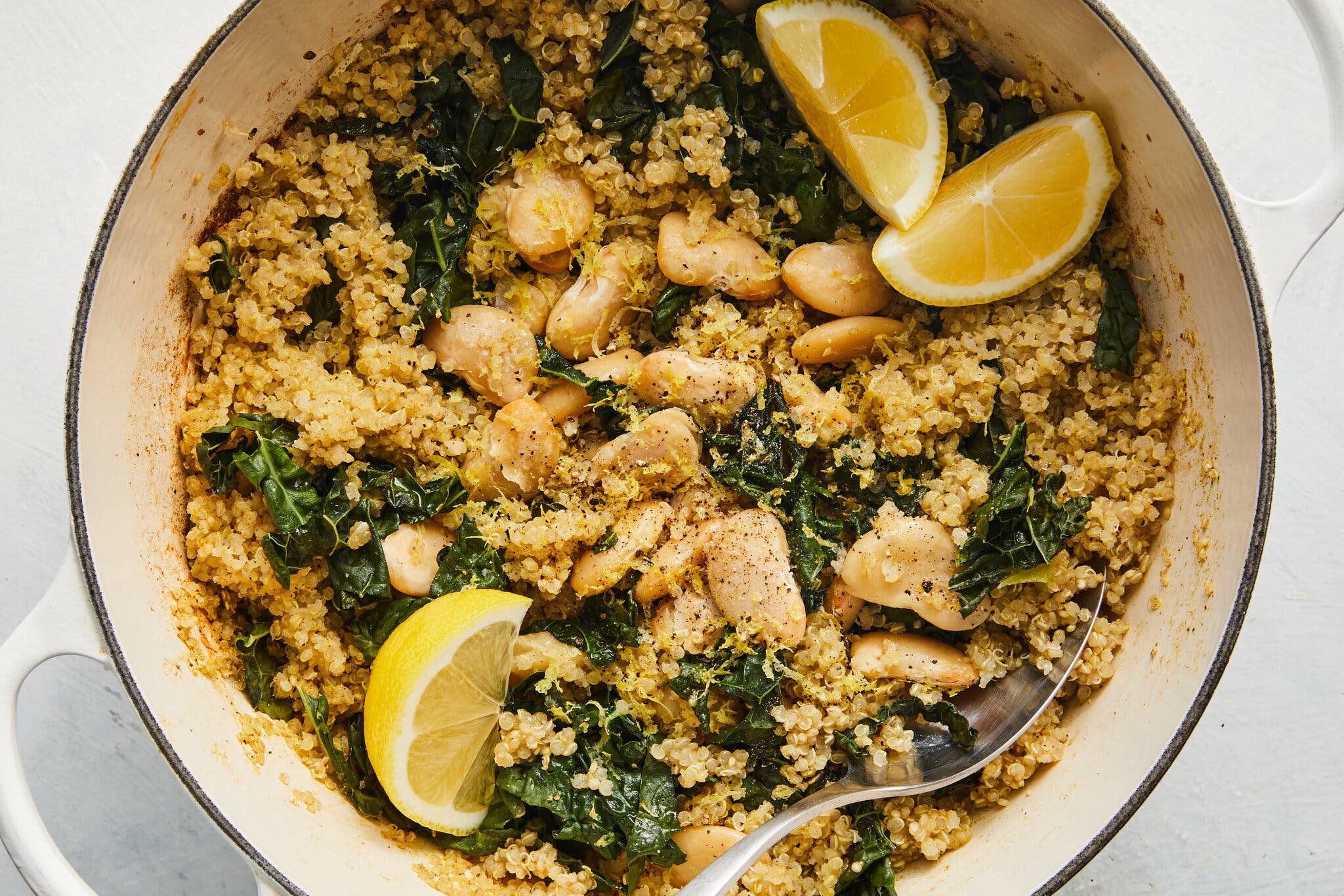 An overhead image of a Dutch oven filled with couscous, greens and white beans. Three lemon wedges sit on top.