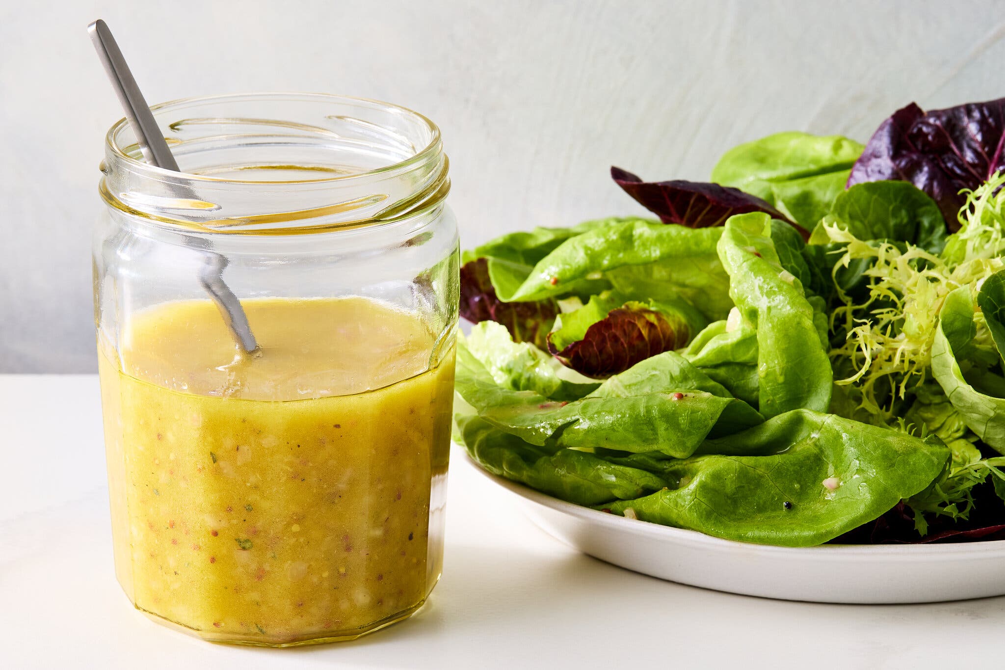 A side image of a clear jar filled with a golden dressing next to a platter of salad greens.