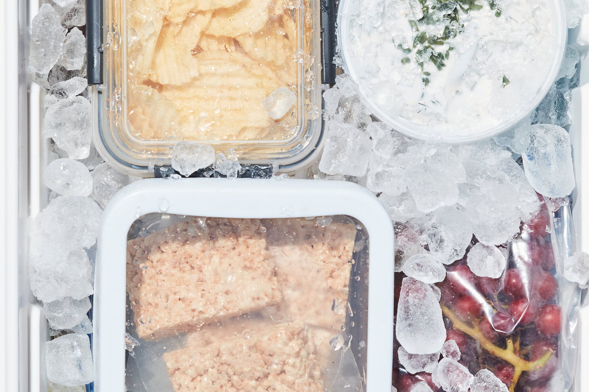 An overhead image of a cooler full of ice and packed with rice krispies treats, chips, dip and grapes.